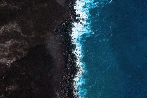 Color pic of aerial photo of a dark gray rocky shoreline on which brilliant blue water breaks into white froth.