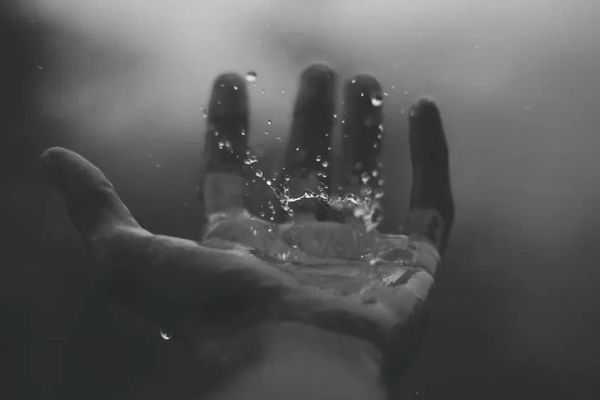 B/W photo of a masculine hand against a blurry background, with a water drop caught mid-splash in the palm