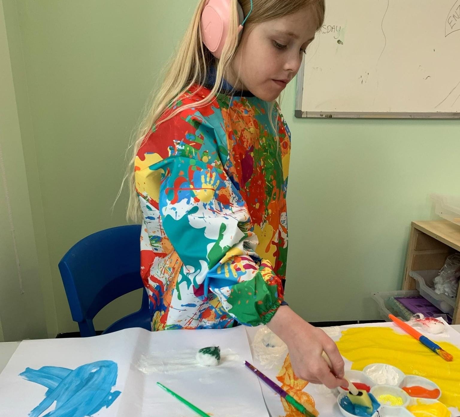 Daisy stands beside a table, dipping a sponge on a stick into some blue paint