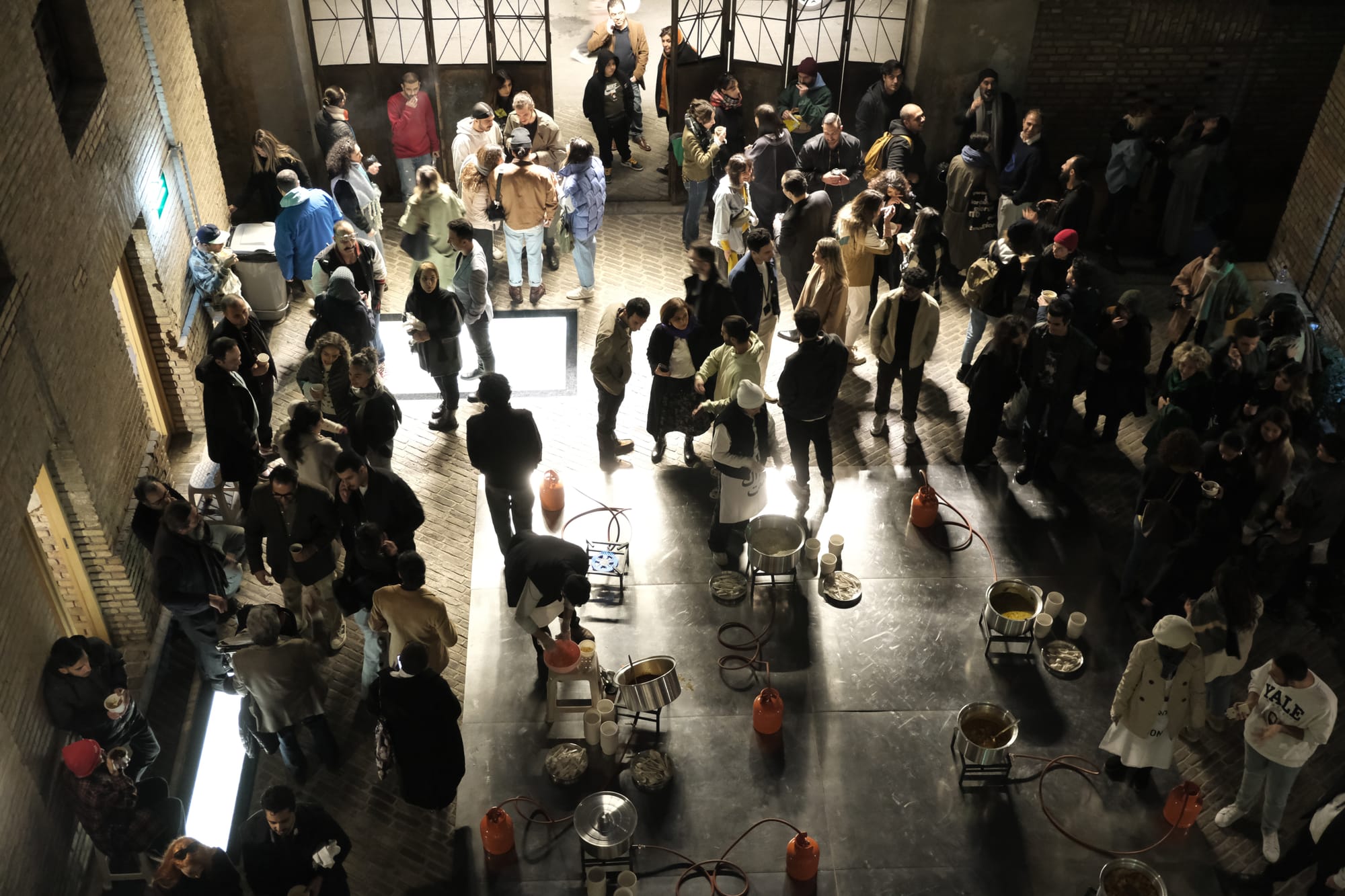 A museum gallery full of people. Stone walls and floors, a few scattered tables. The doors to the outside are open.