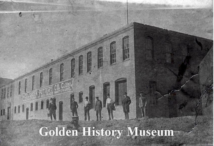 Two story brick industrial building with Geijsbeek Pottery CO painted on the side.  Seven men are standing front of the building, facing the camera.