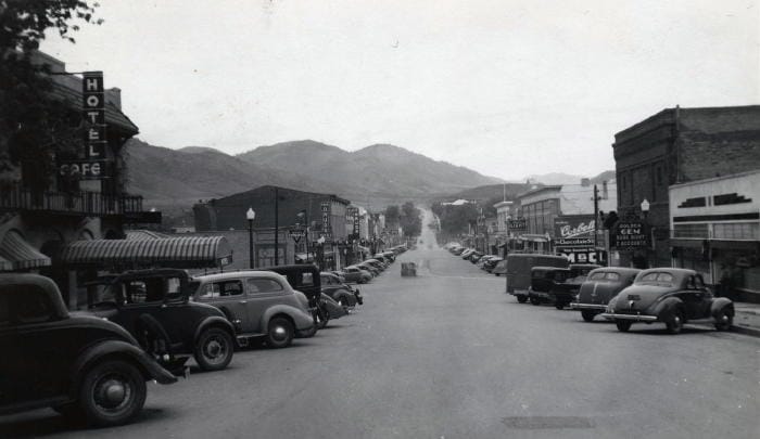 Black and white photo showing the commercial district of Washington Avenue in Golden Colorado. Cars at diagonally parked on both sides of the street. Many signs jut out from the buildings, including Hotel/Cafe, Conoco, Foss Drugs, Alpine Drugs, Golden Gem, Chocolate Shop, and Central Light and Power