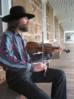 Bearded man in a cowboy hat sits on a bench in front of a stone building, playing a violin.