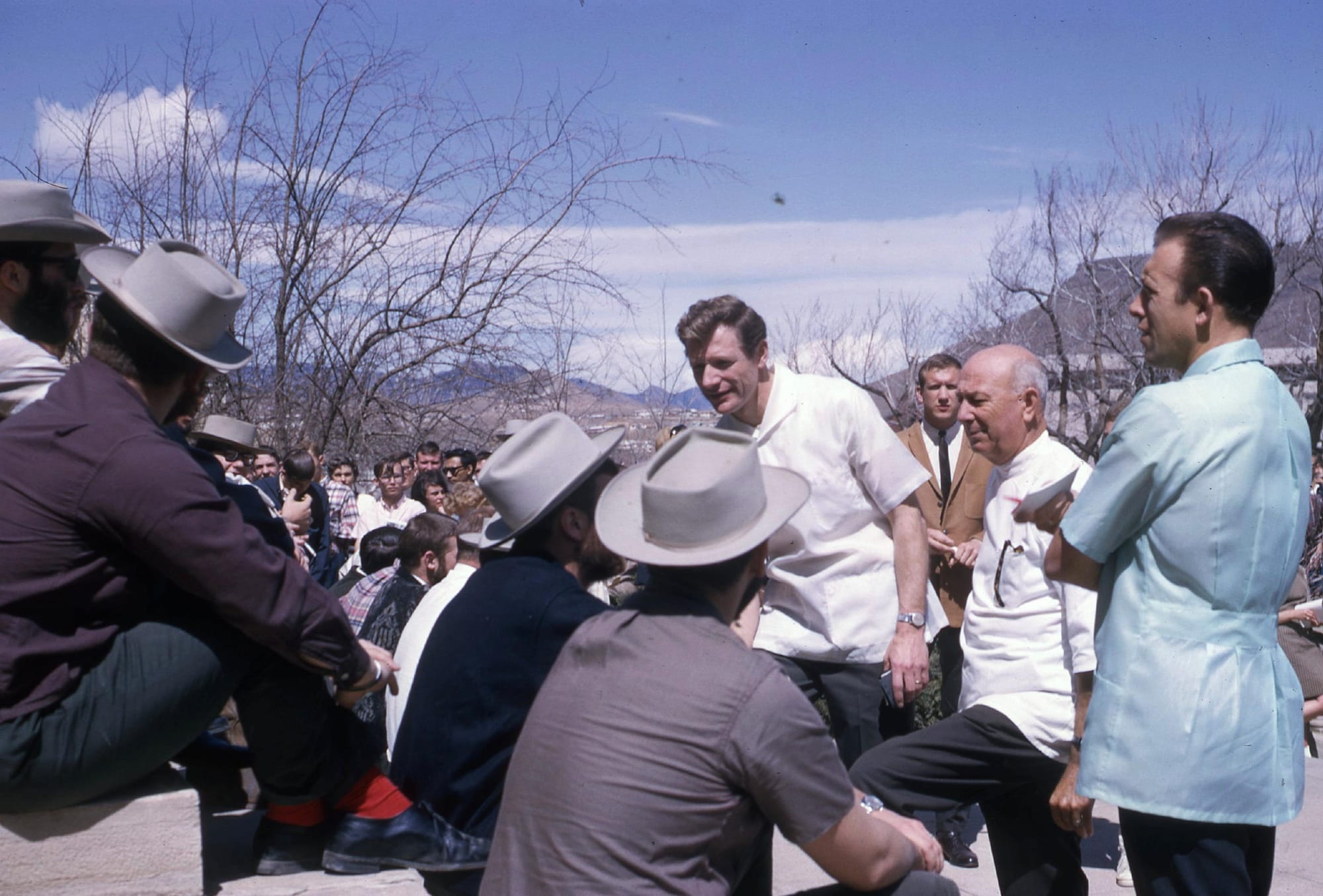 Several men wearing gray hats sit on bleachers.  Three men in barber's smocks examine them.