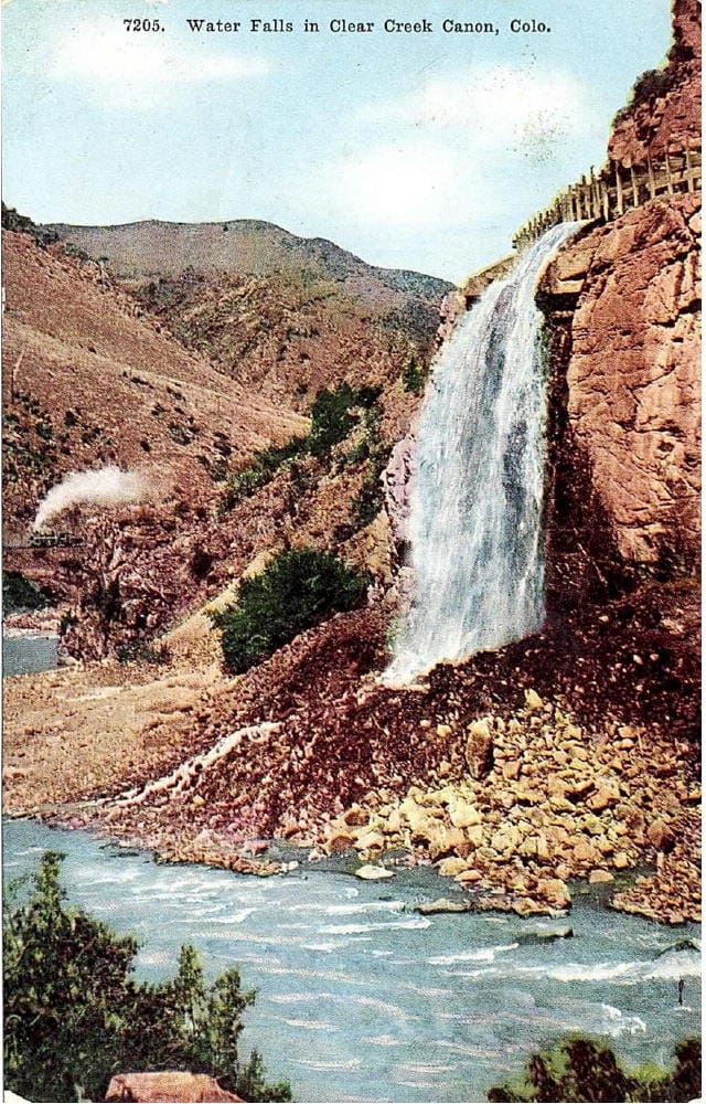 A wooden flume high over the creek creates a waterfall tumbling down.  Steam engine in the distance.