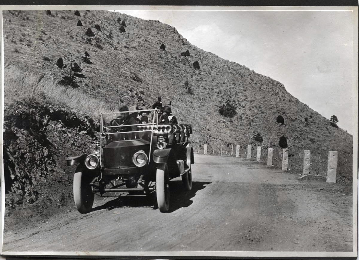 Open air touring car with several rows of seats and a few passengers on a winding gravel mountain road.