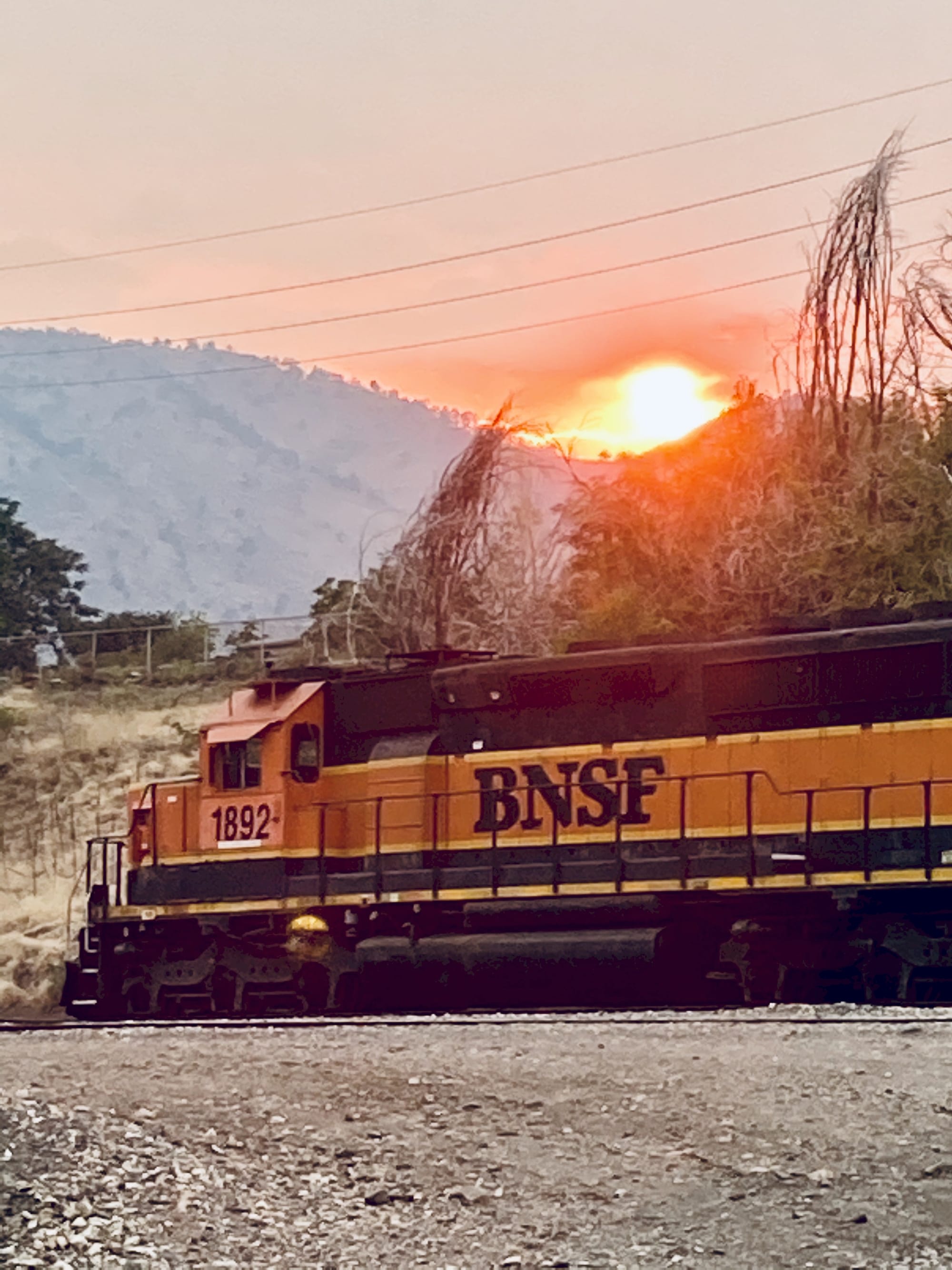 Orange and black locomotive under a bright orange and yellow sunset surrounded by red clouds