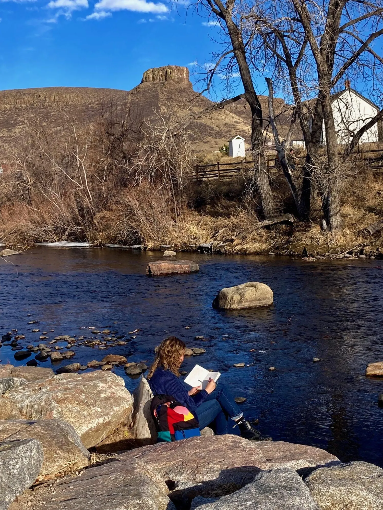 Young woman sitting on rocks next to Clear Creek, holding a book and a pen.