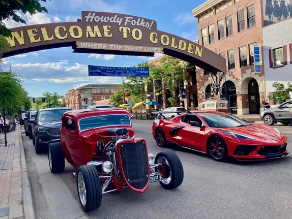 two red cars under the downtown welcome arch--a sports car in the driving lane and a Roadster parked on the side