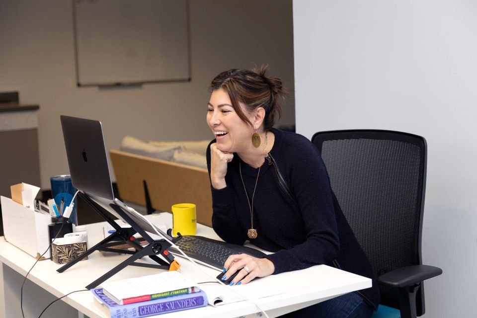 a woman wearing a black sweater with long hair pulled back is sitting at a desk and laughing at something on a laptop screen