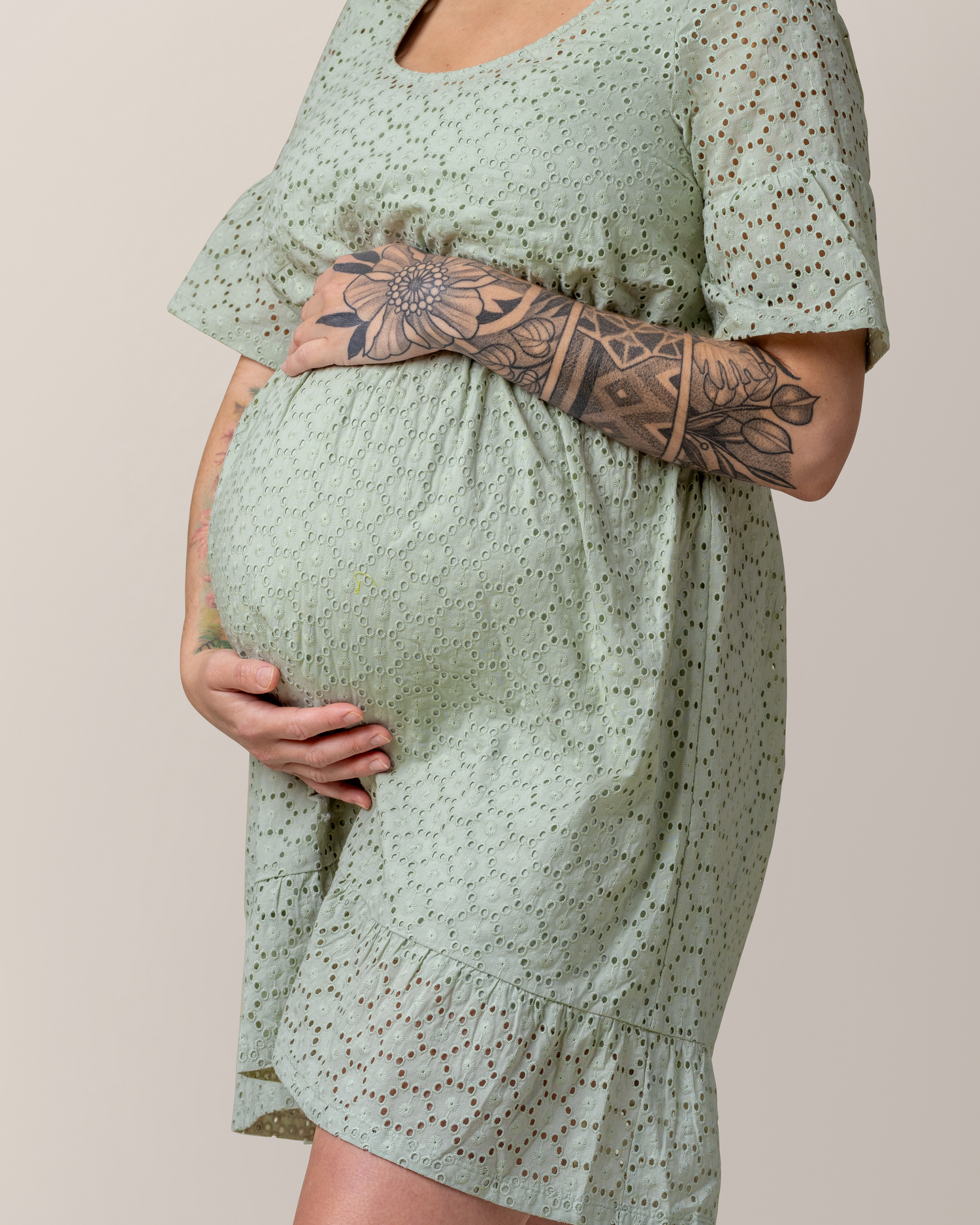 Pregnant woman in a sage green eyelet dress holding her baby bump with both hands, showcasing a detailed floral tattoo on her forearm.