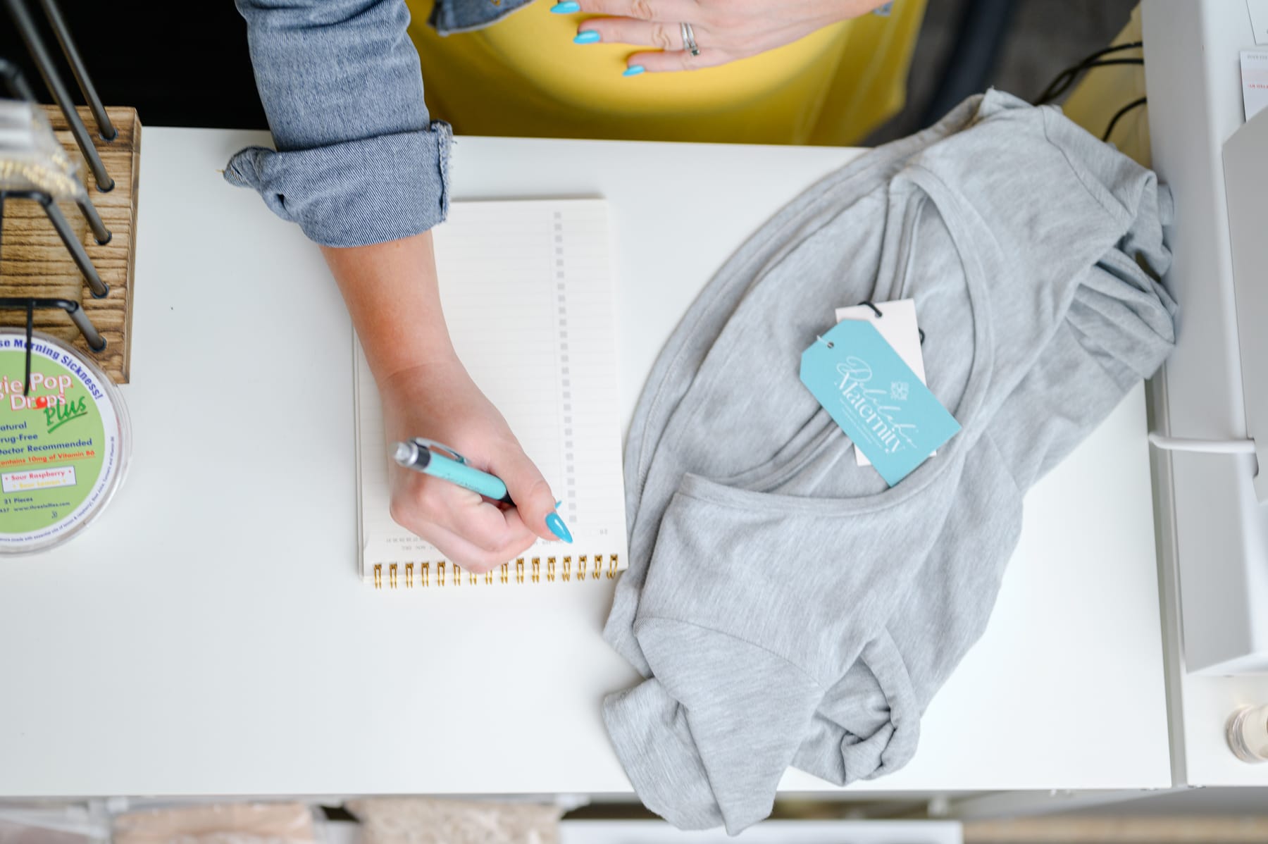 Person writing in a spiral notebook beside a folded gray maternity top with a Delilah's Maternity tag, hinting at planning or shopping for gifts.