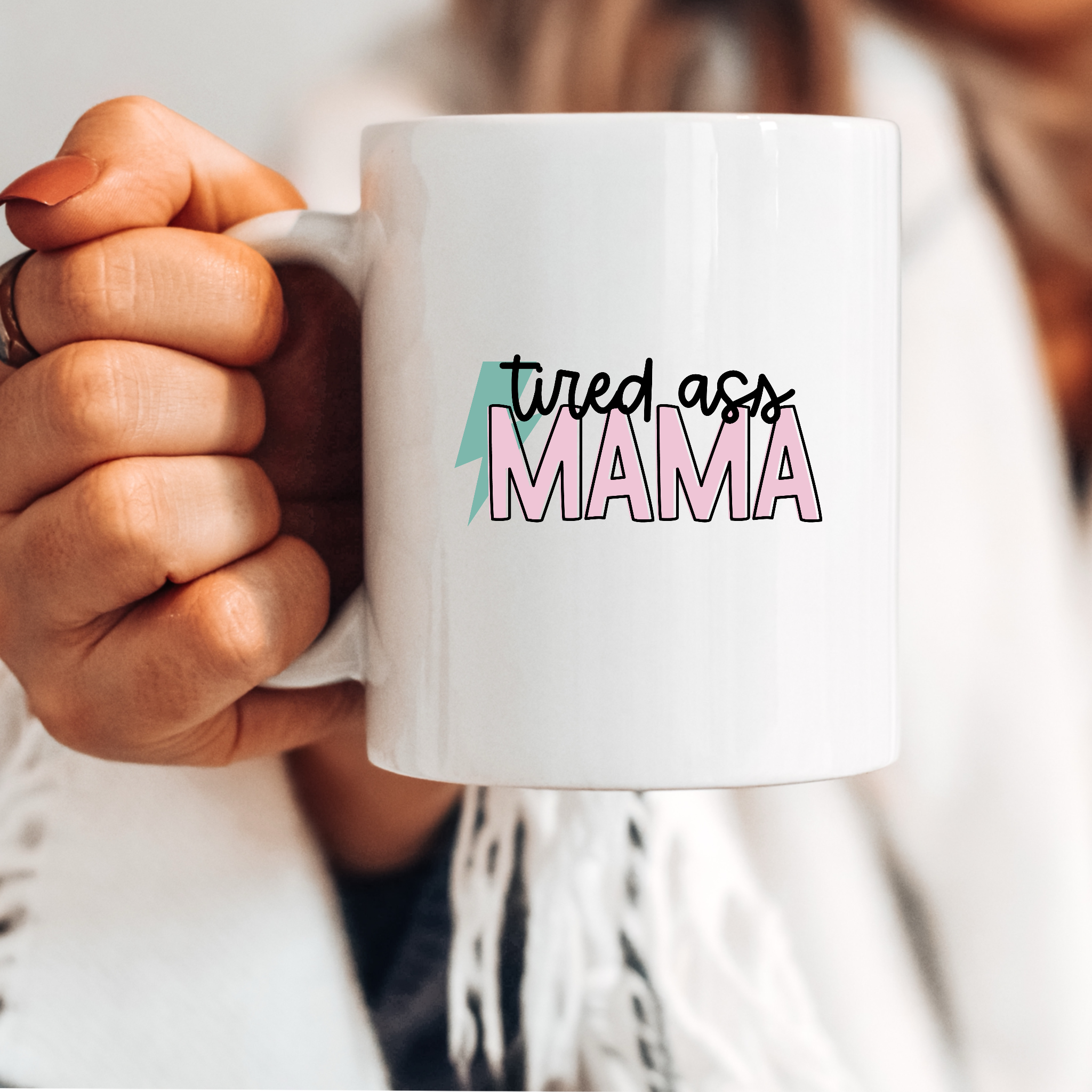 Close-up of a hand holding a white coffee mug with the playful phrase 'Tired Ass Mama' printed in pink and black lettering.