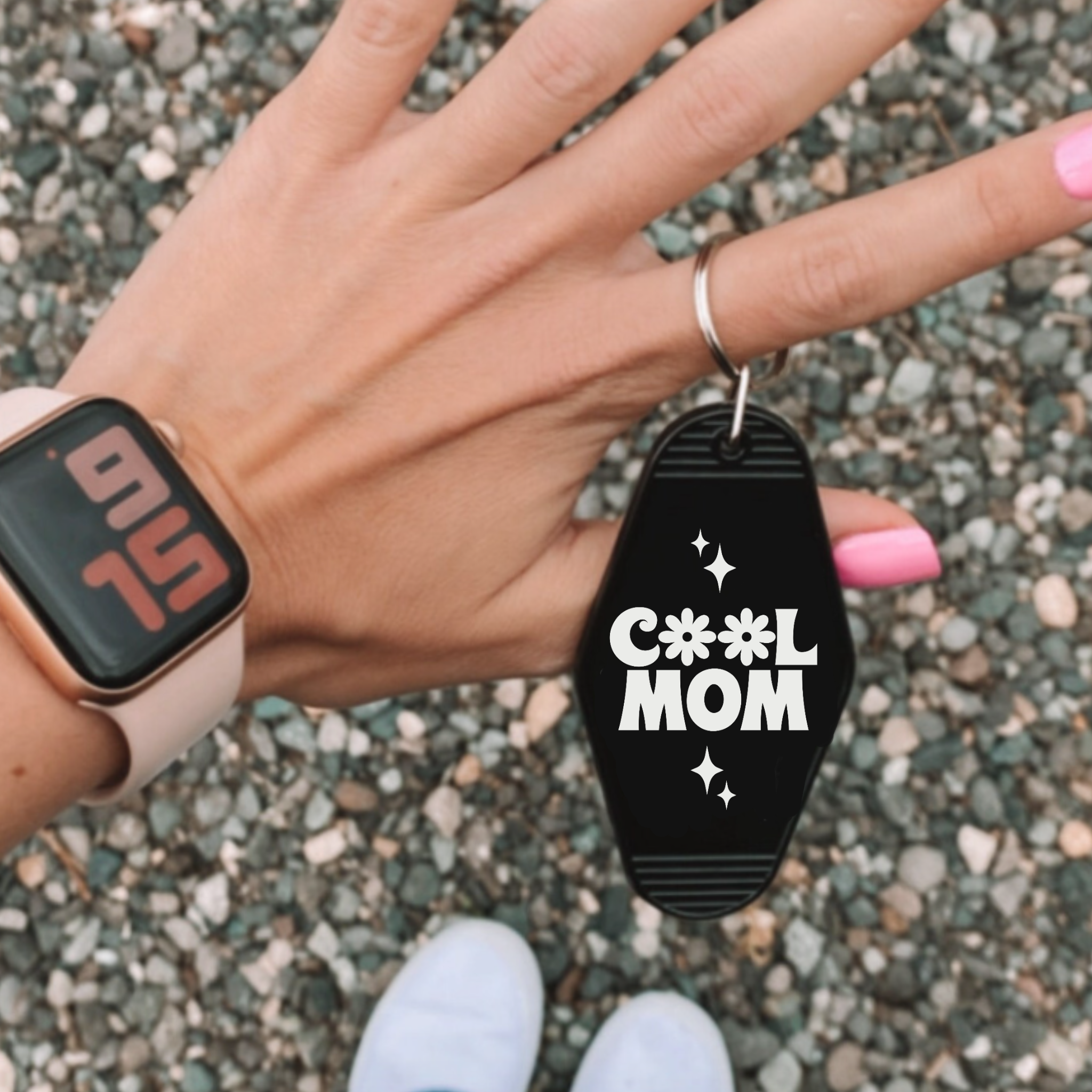 Hand holding a black retro-style keychain that reads 'Cool Mom' in bold white letters, with a pink smartwatch and pink-painted nails in view.