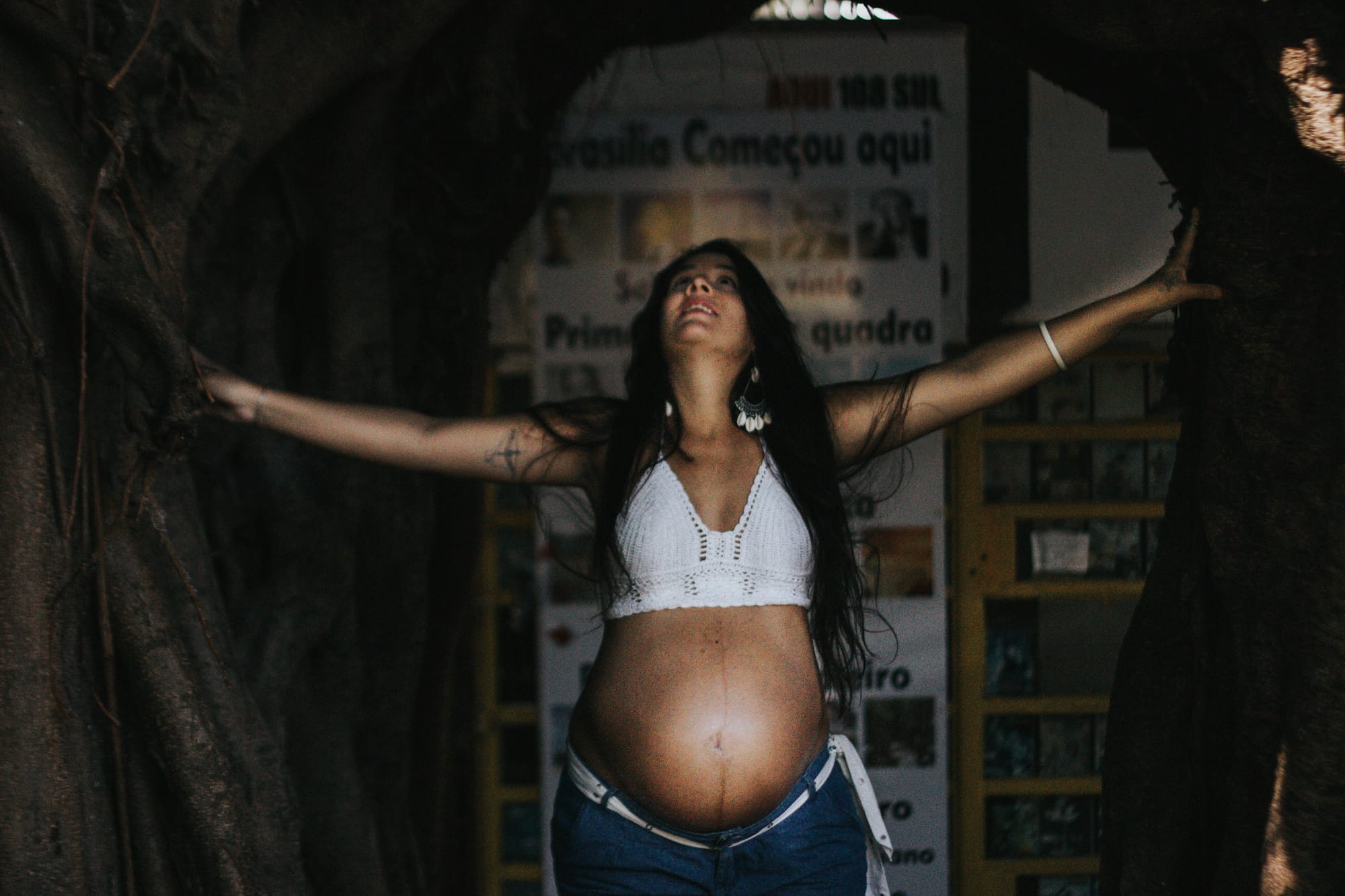 A woman in a white bralette top and jeans showing her bare, pregnant belly while smiling.