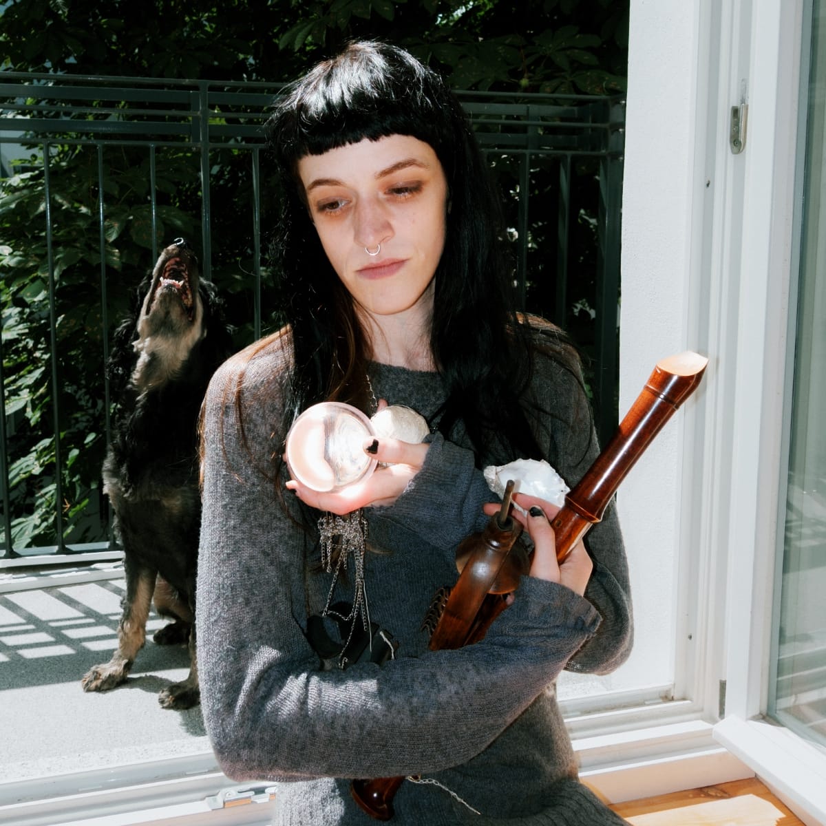 Bridget Ferrill seated, holding a clock and a woodwind instrument as a dog barks in the background. Photo by Elizabeth Davis.