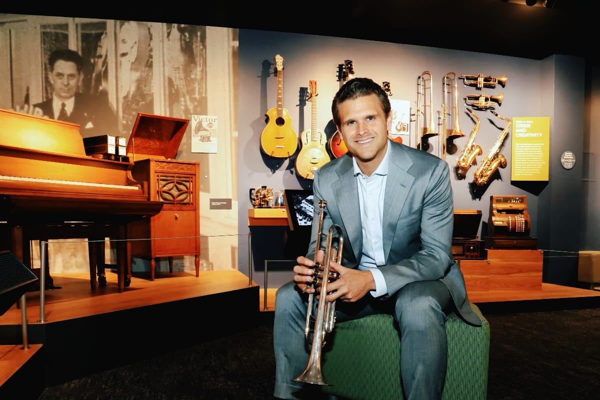 John Mlynczak sitting, surrounded by instruments, holding a trumpet