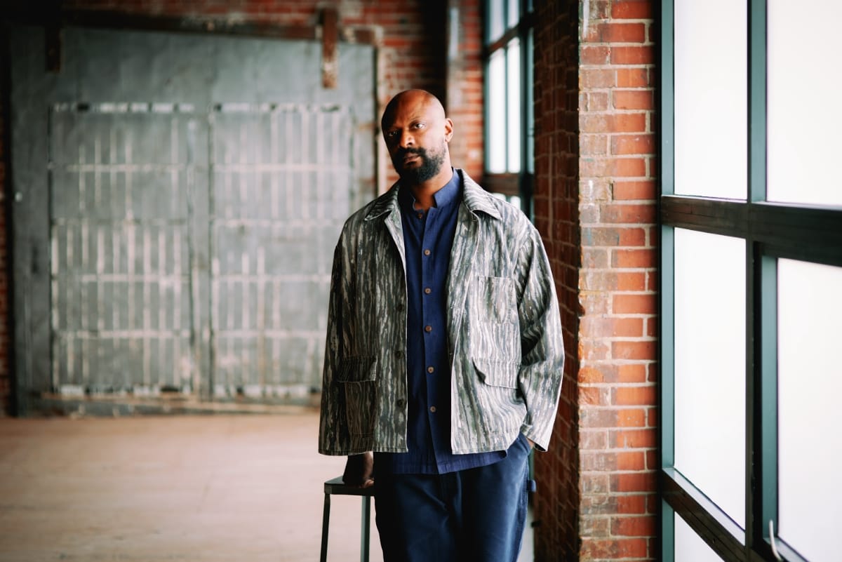 Justin Hicks in patterned jacket and blue shirt stands in industrial loft space with exposed brick and large windows. Photo by Michael Bueno.