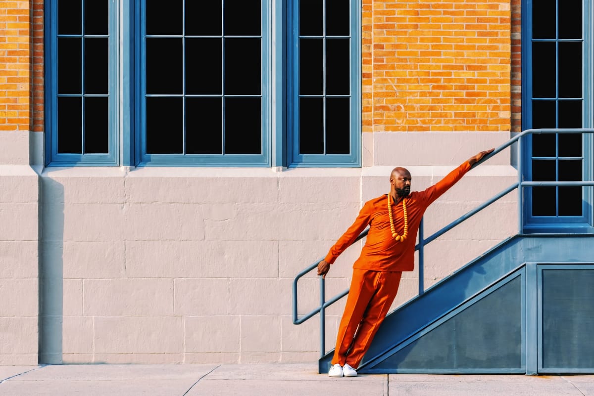 Justin Hicks in orange outfit with wooden beads on concrete stairs, blue-framed windows and orange brick backdrop. Photo by Malik Welli.