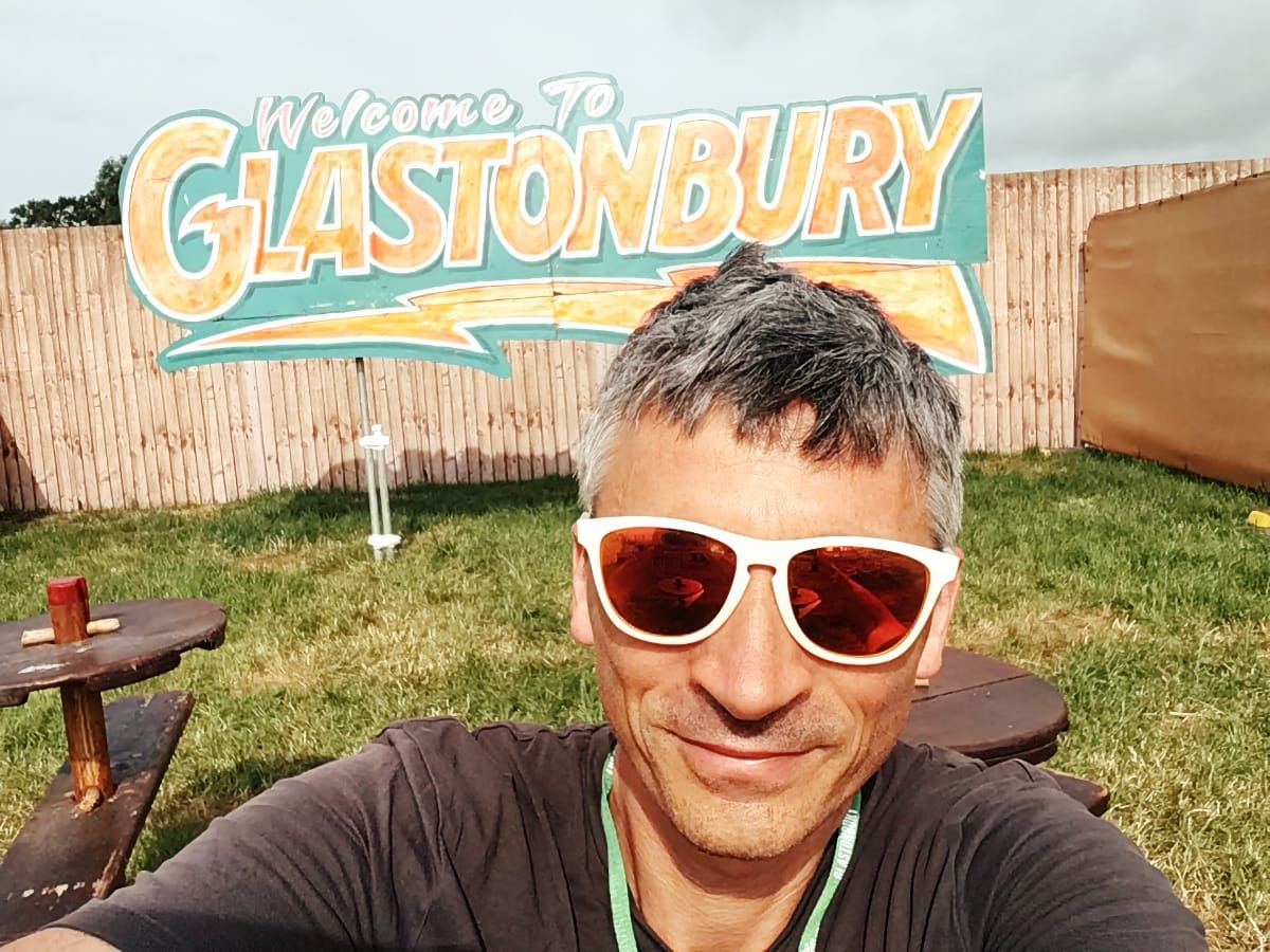 Ludo Hunter-Tilney in white sunglasses takes a selfie at Glastonbury Festival with the colorful welcome sign behind him.