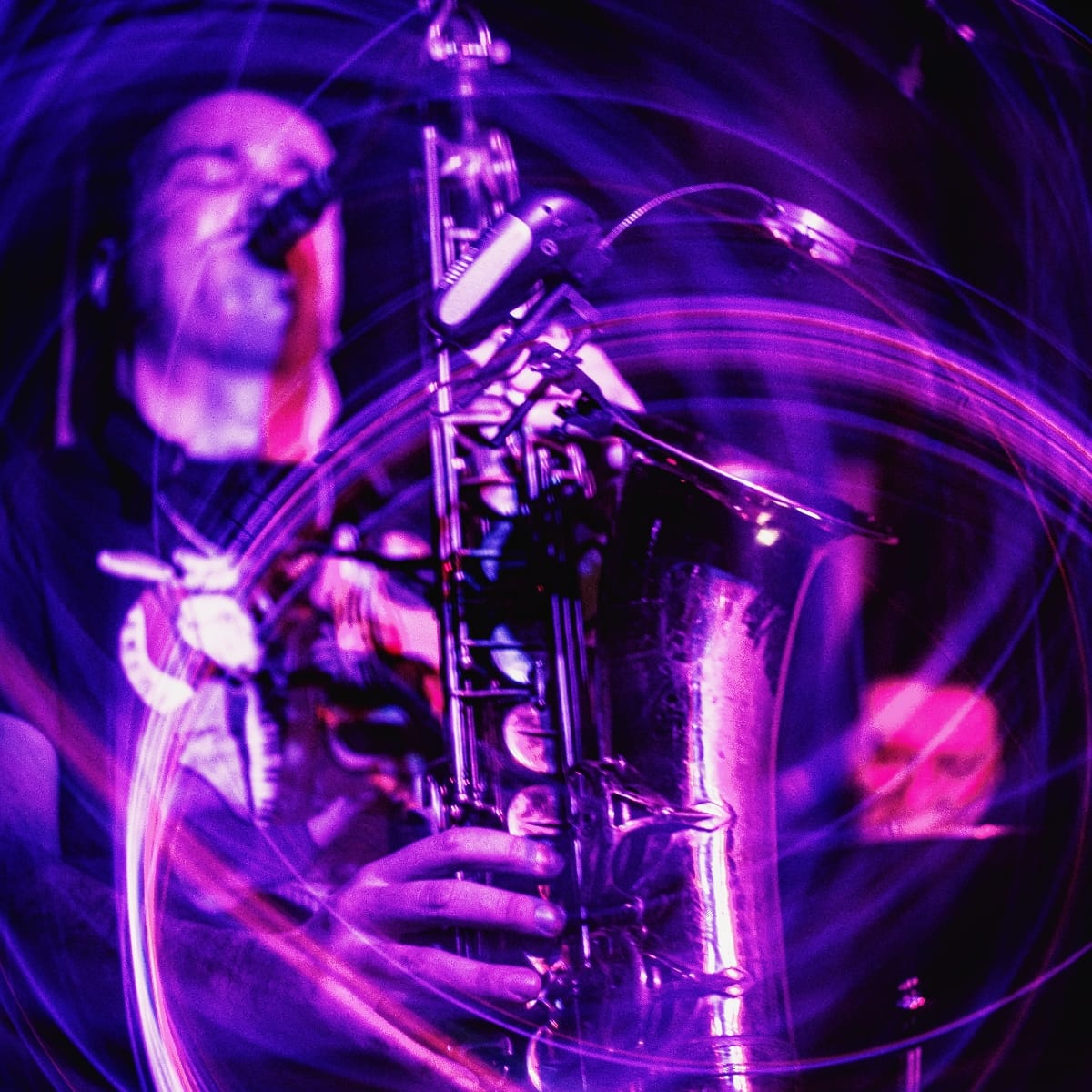 Patrick Smith plays saxophone captured through distorted lens effect, bathed in vivid purple and magenta stage lighting. Photo by Camille Neirynck-Guerrero.