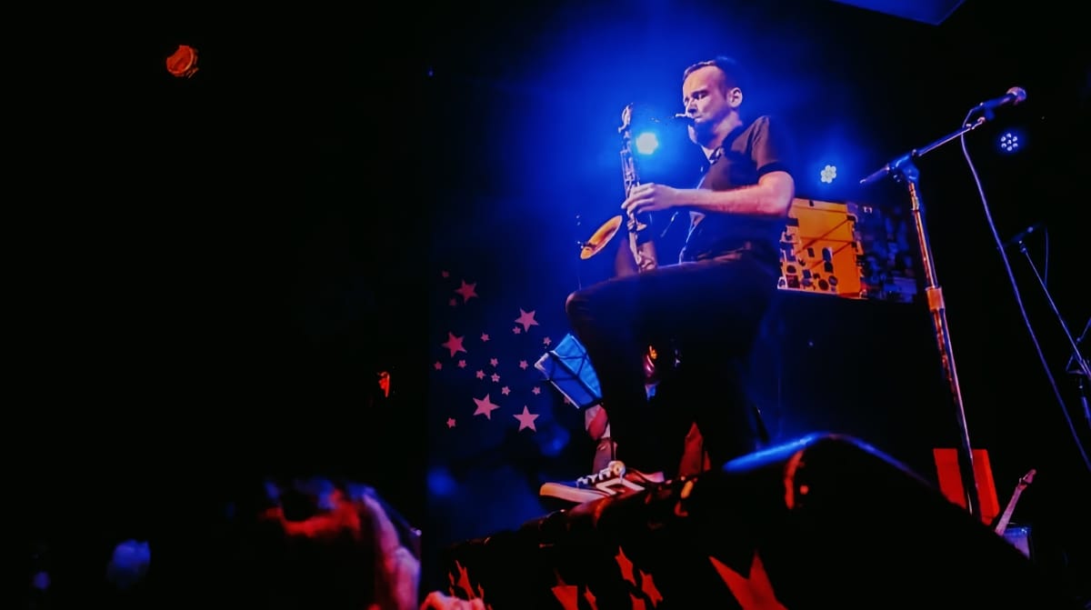 Patrick Smith performs on saxophone under blue stage lighting with decorative stars and props visible, audience in foreground.