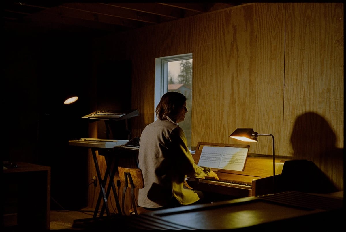 Erik Hall at piano in wood-paneled studio, natural light through window, music sheets illuminated by desk lamp, warm moody atmosphere. Photo by Nolis Anderson.