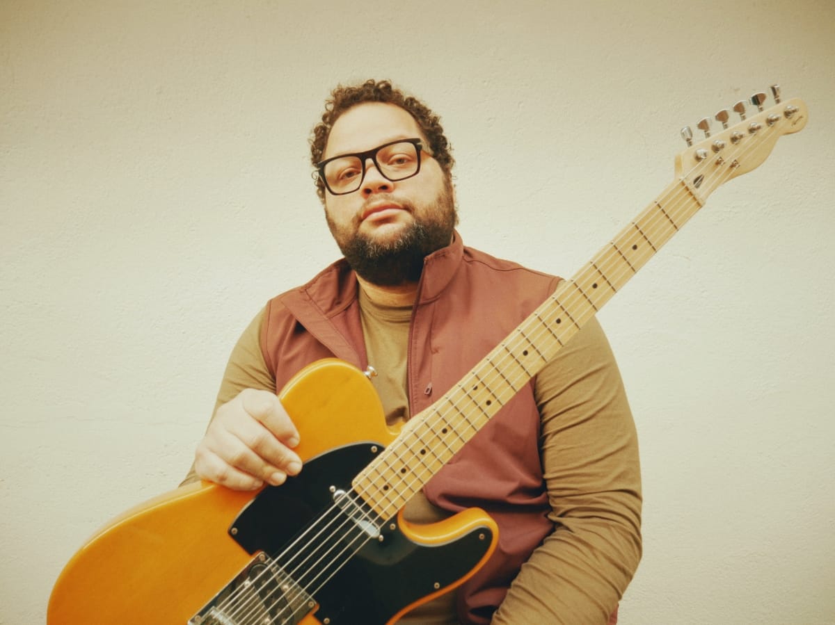 Juanma Trujillo holds yellow electric guitar, wearing earth-toned clothing and glasses against plain wall, warm vintage tones. Photo by Leonor Falcón.