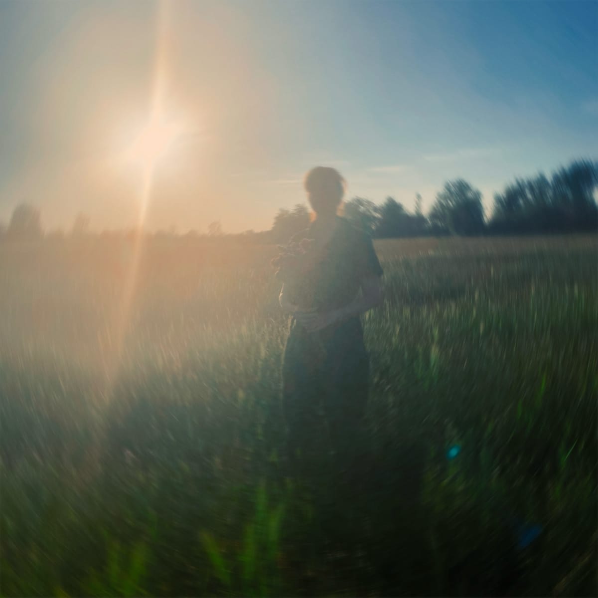 Loula Yorke in motion-blurred field at golden hour, creating a soft, dreamy atmosphere with sun flare and distant trees.