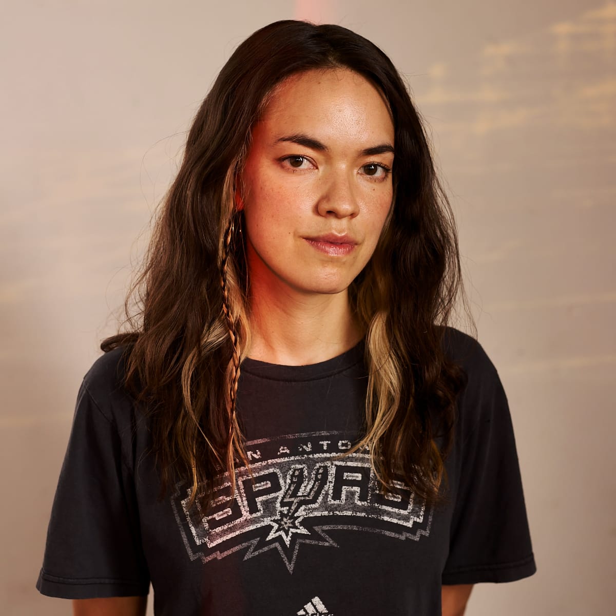 Momoko Gill in San Antonio Spurs t-shirt with long wavy hair, small braid, and hoop earrings against neutral backdrop. Photo by Manuel Vasquez.