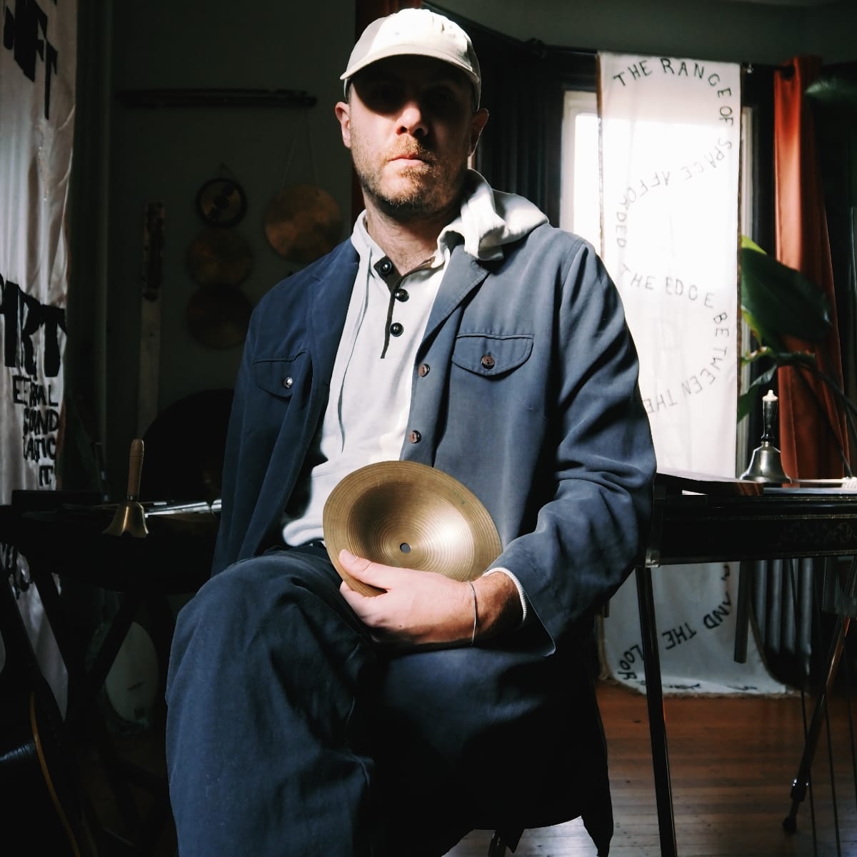 Sam Wenc in white cap and blue jacket holds brass cymbal in home studio, text banners visible behind.