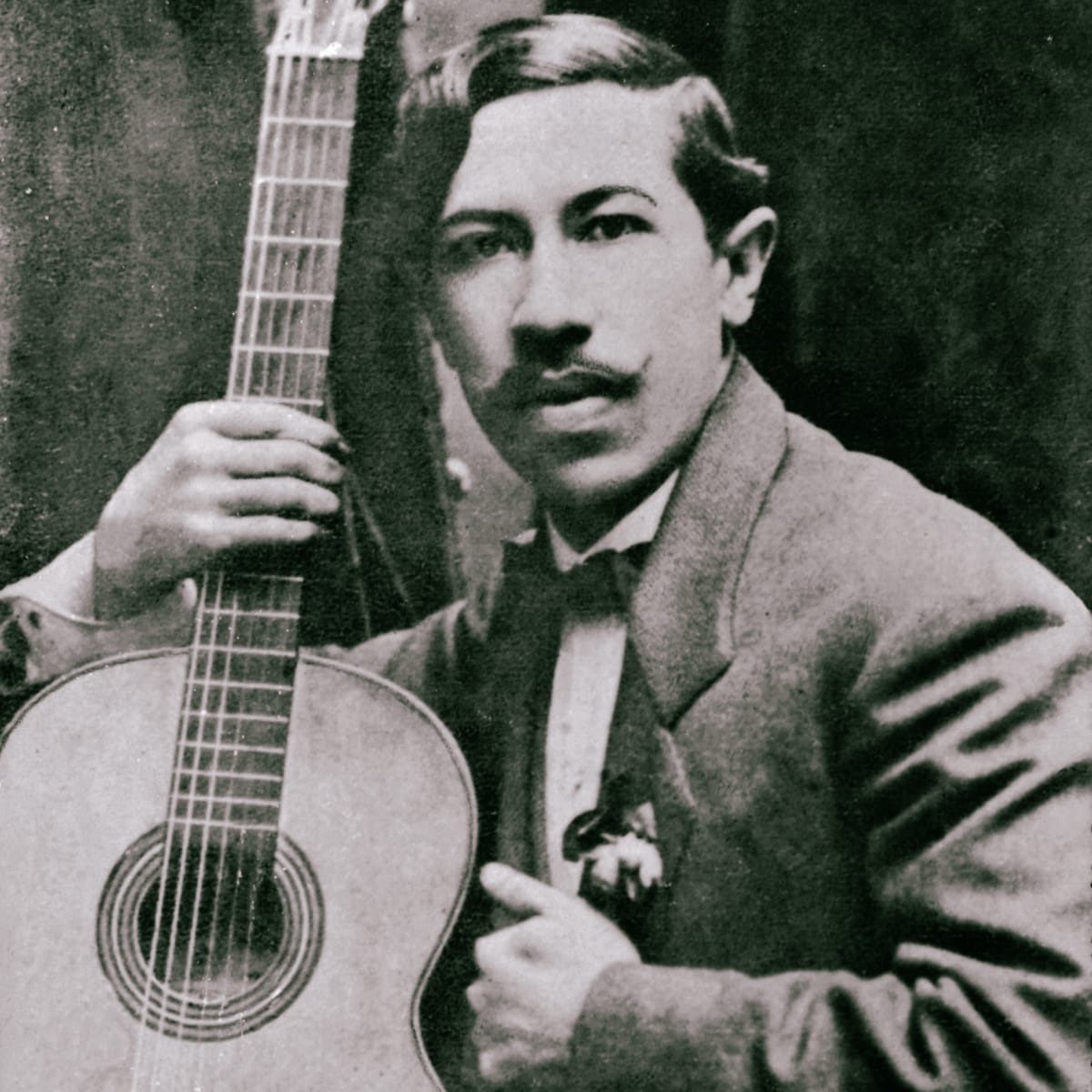 A sepia studio portrait of a young Agustín Barrios-Mangoré in a suit and boutonniere, holding a classical guitar upright beside him, circa 1910.