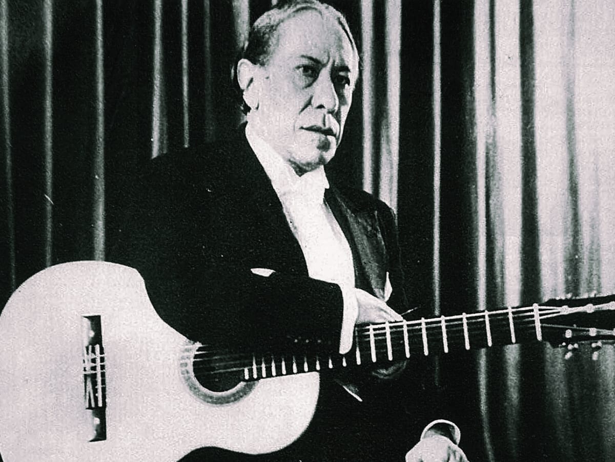 A black-and-white photo of an older Agustín Barrios-Mangoré seated in a formal suit with a classical guitar, against a draped curtain backdrop, 1940s.