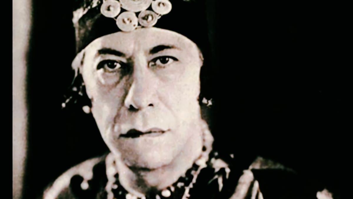 A close-up film still of Agustín Barrios-Mangoré in his Nitsuga Mangoré persona, wearing an ornate indigenous headdress and beaded costume against a dark background.