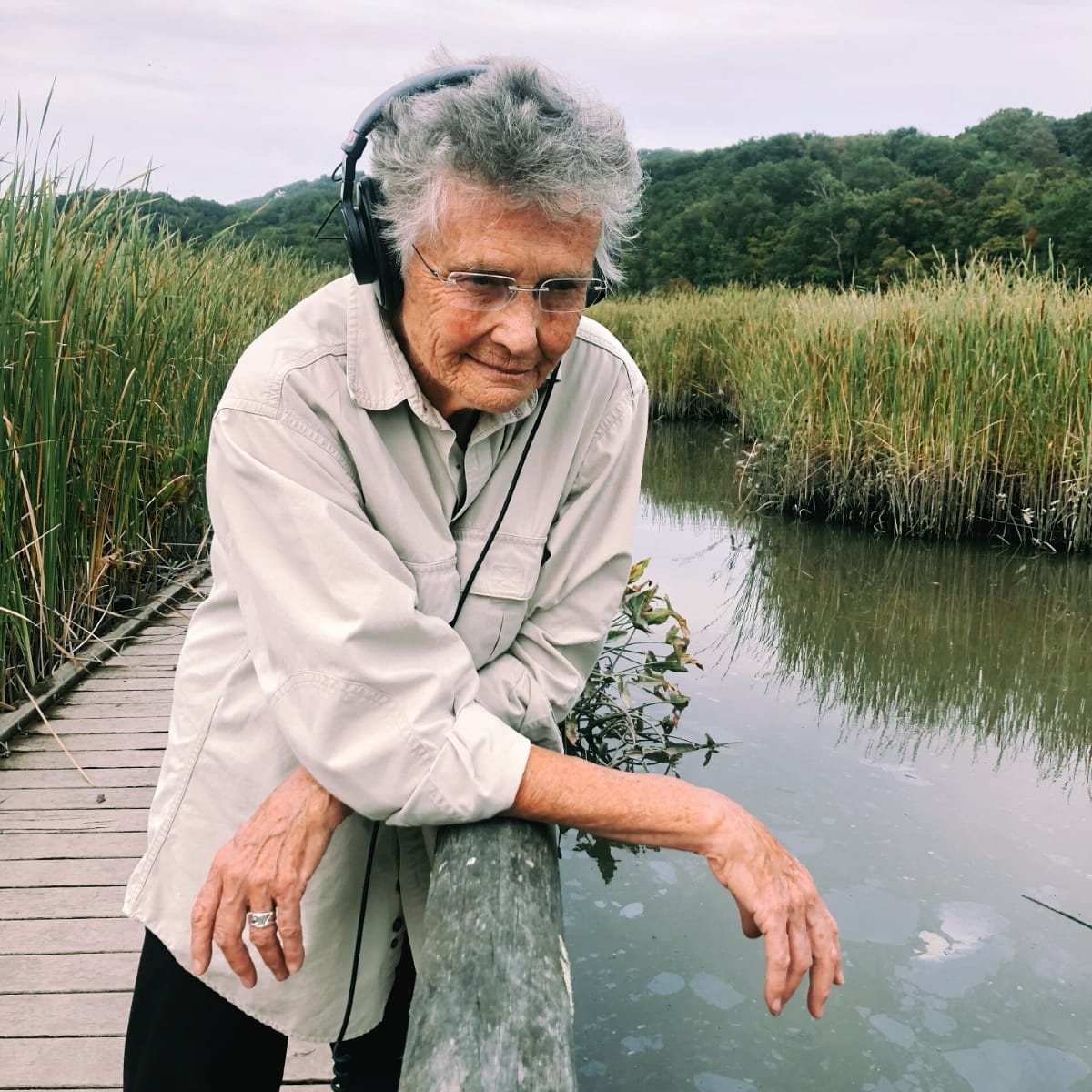 Annea Lockwood leans over a wooden dock railing wearing headphones, gazing at the water of Constitution Marsh, surrounded by tall reeds. Photo by Sam Green / Free History Project.