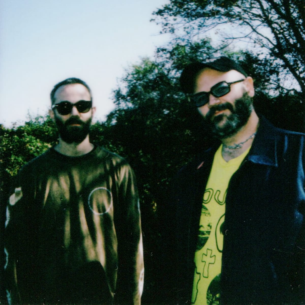 A Polaroid-style outdoor photo of John Thayer and Ben Seretan, both bearded and wearing sunglasses, one in a metallic shirt and one in a yellow graphic tee and jacket.