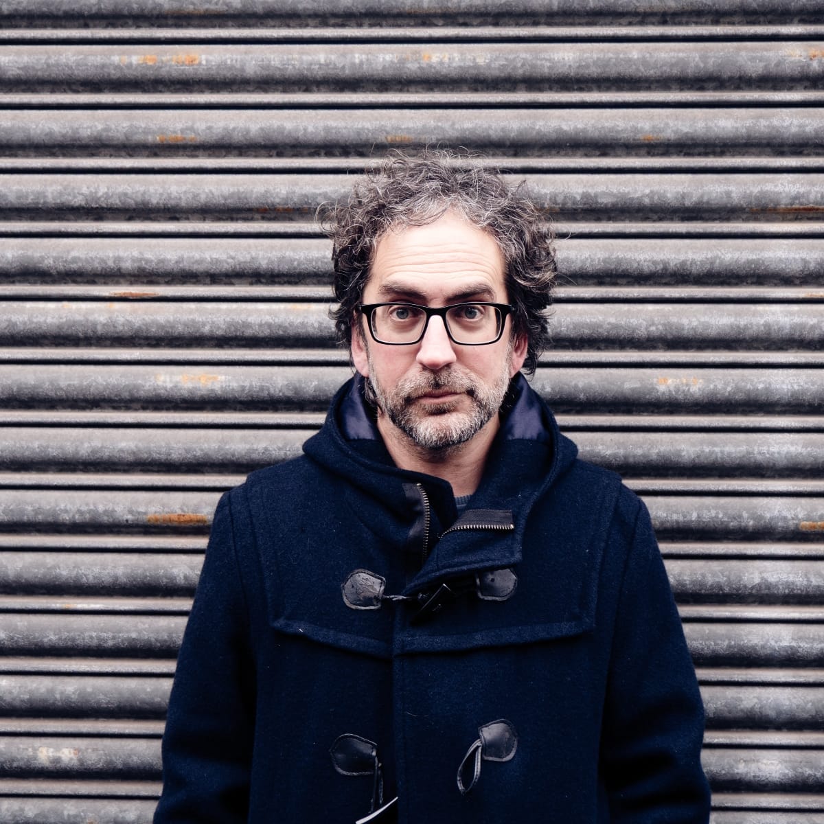 Billy Fuller stands in a dark duffle coat before a corrugated metal shutter, looking directly at the camera. Photo by Charlie Romijn-Barr.