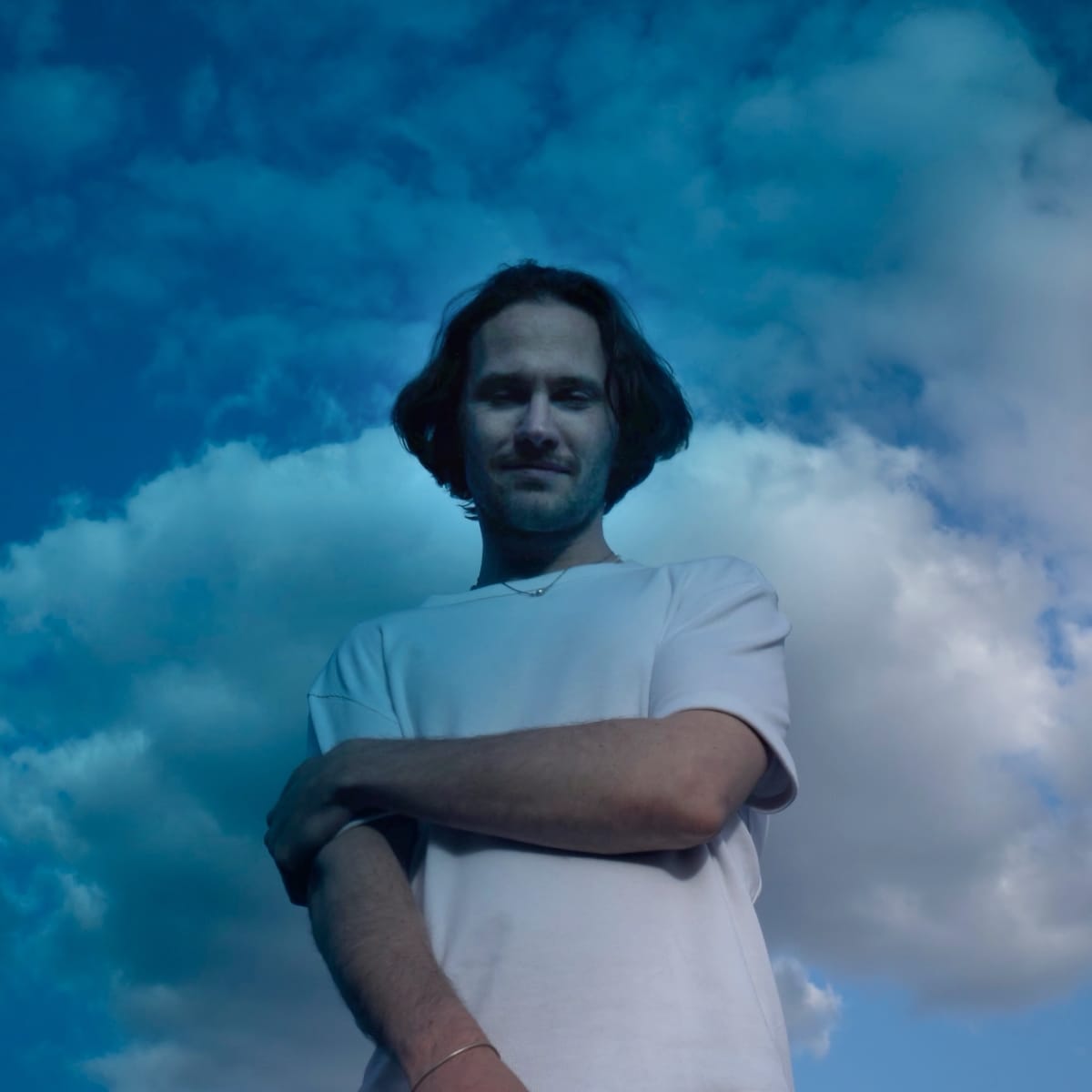 A man (David August) with shoulder-length hair and a white t-shirt stands arms crossed against a vivid blue sky with dramatic clouds. Photo by Sarah Fuchs.