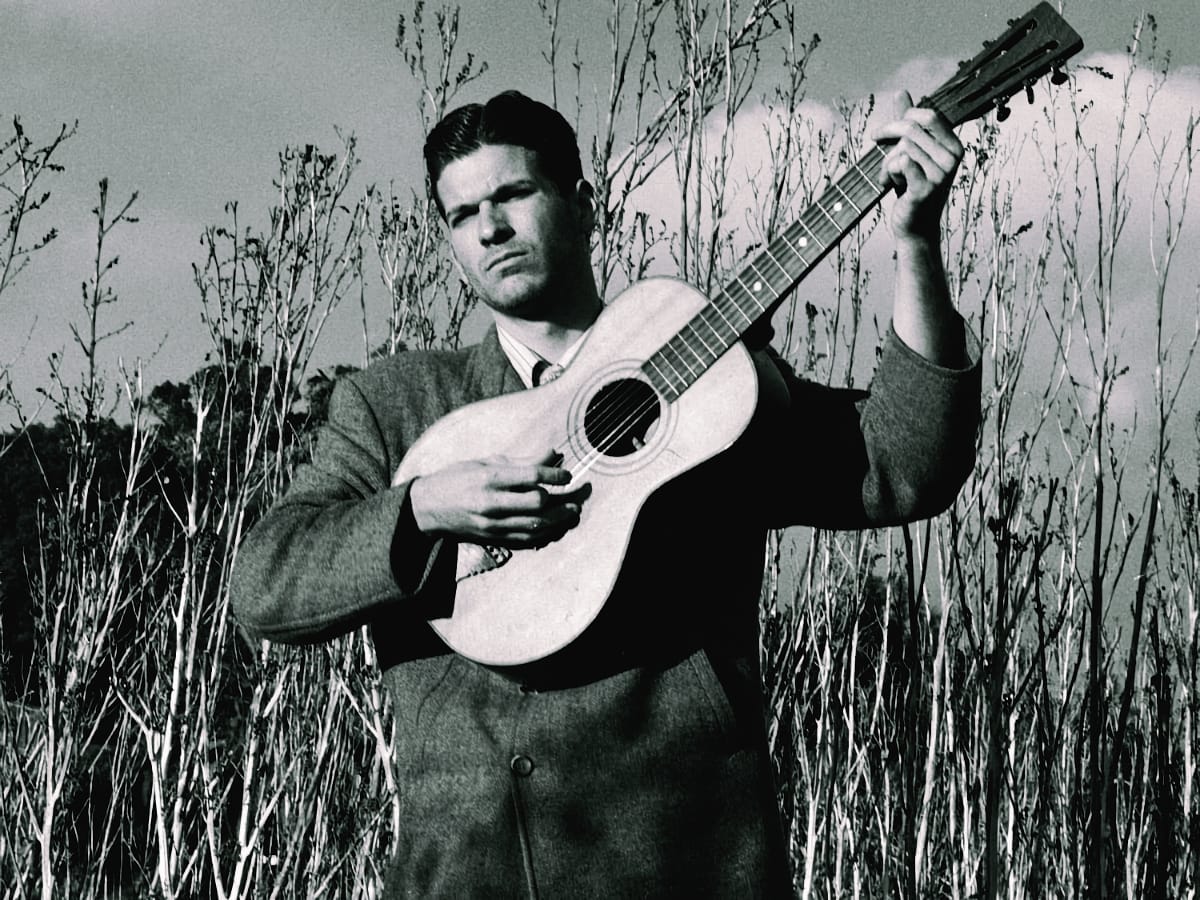 A black-and-white photo of Frank Fairfield playing an acoustic guitar while standing among tall dry brush beneath an overcast sky.