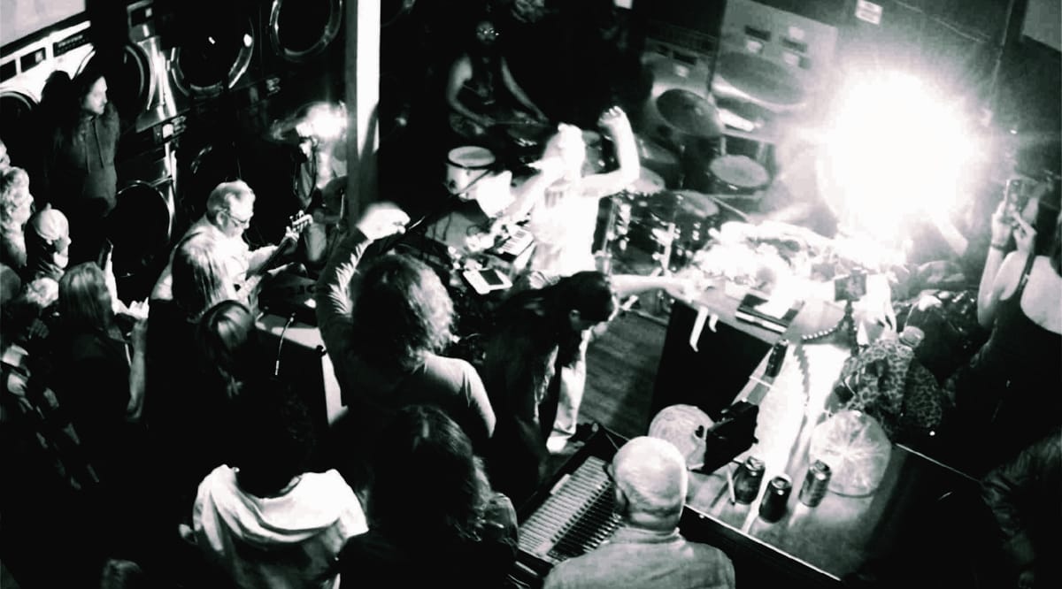 Galecstasy and Mike Watt Trio perform in a laundromat, shot from above; a bassist, vocalist, and drummer play amid a dense surrounding crowd, washers lining the walls.  Photo by Suraj Patra.