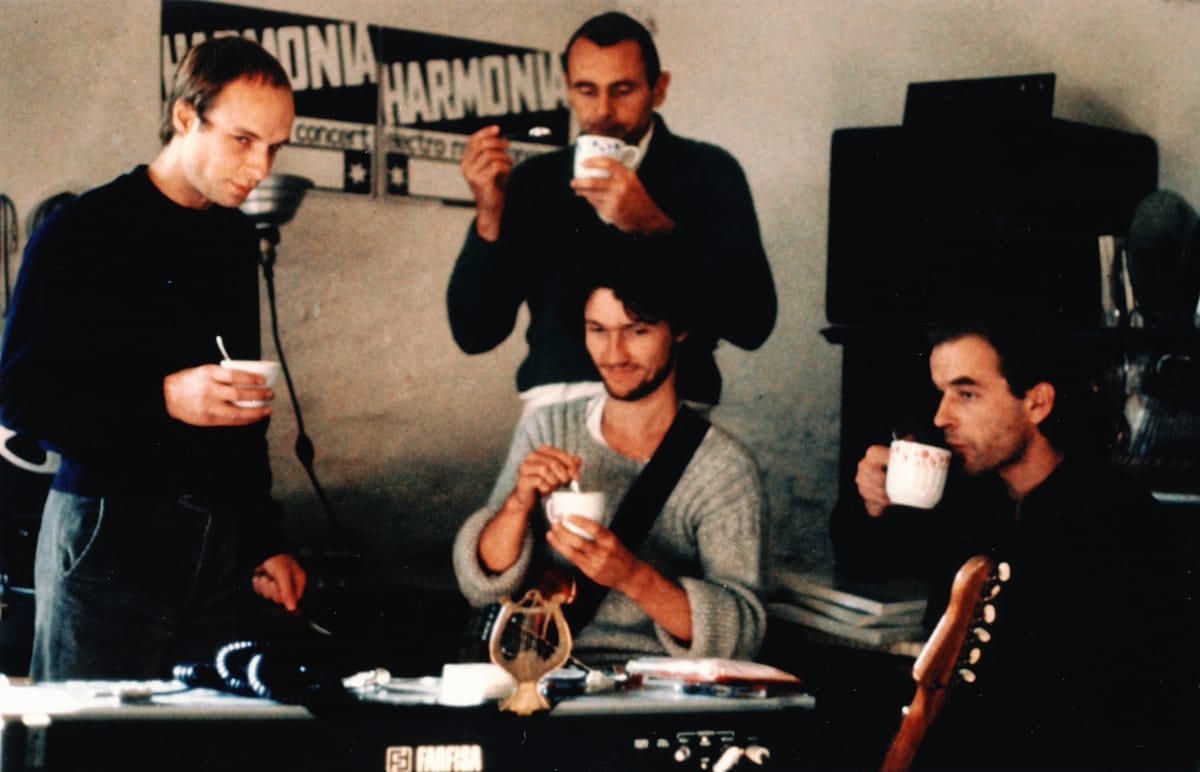 A vintage photo of Brian Eno, Hans-Joachim Roedelius, Michael Rother, and Dieter Moebius sharing coffee or tea in a room with Harmonia concert banners on the wall behind them.
