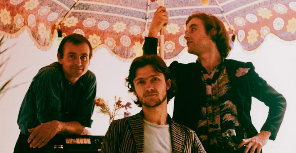 A vintage color photo of Hans-Joachim Roedelius, Michael Rother, and Dieter Moebius of Harmonia grouped beneath a patterned parasol, with a small keyboard and flowers visible behind them.