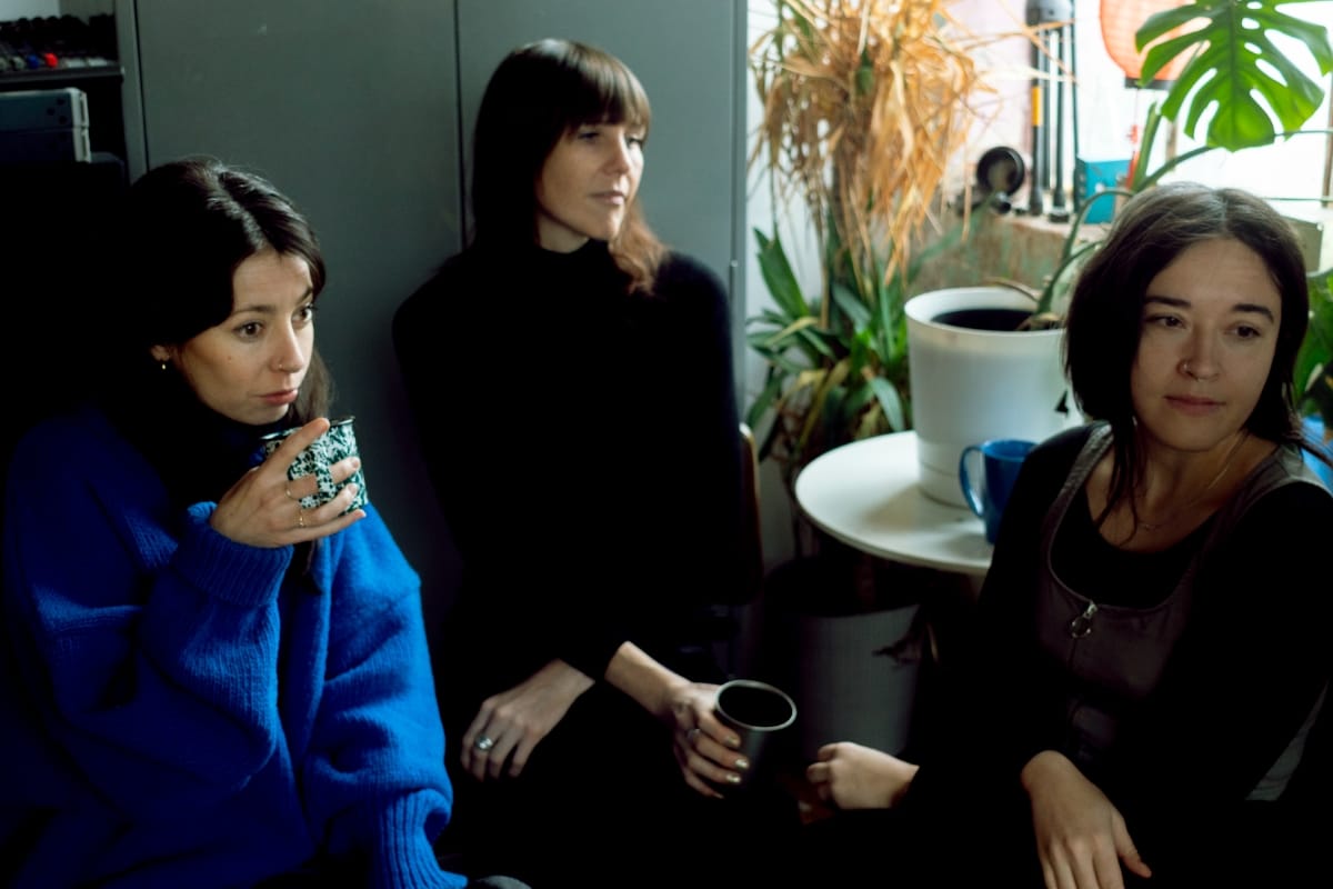 Lia Kohl, Whitney Johnson, and Macie Stewart sit together in a plant-filled room, two holding mugs, one in a bright blue sweater and the others in black. Photo by Jack Garland.