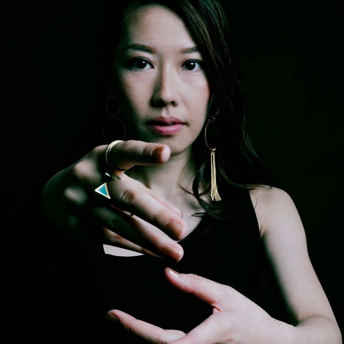 Miho Hazama extends a ringed hand toward the camera, wearing gold tassel earrings and a black top, lit dramatically against a black background.