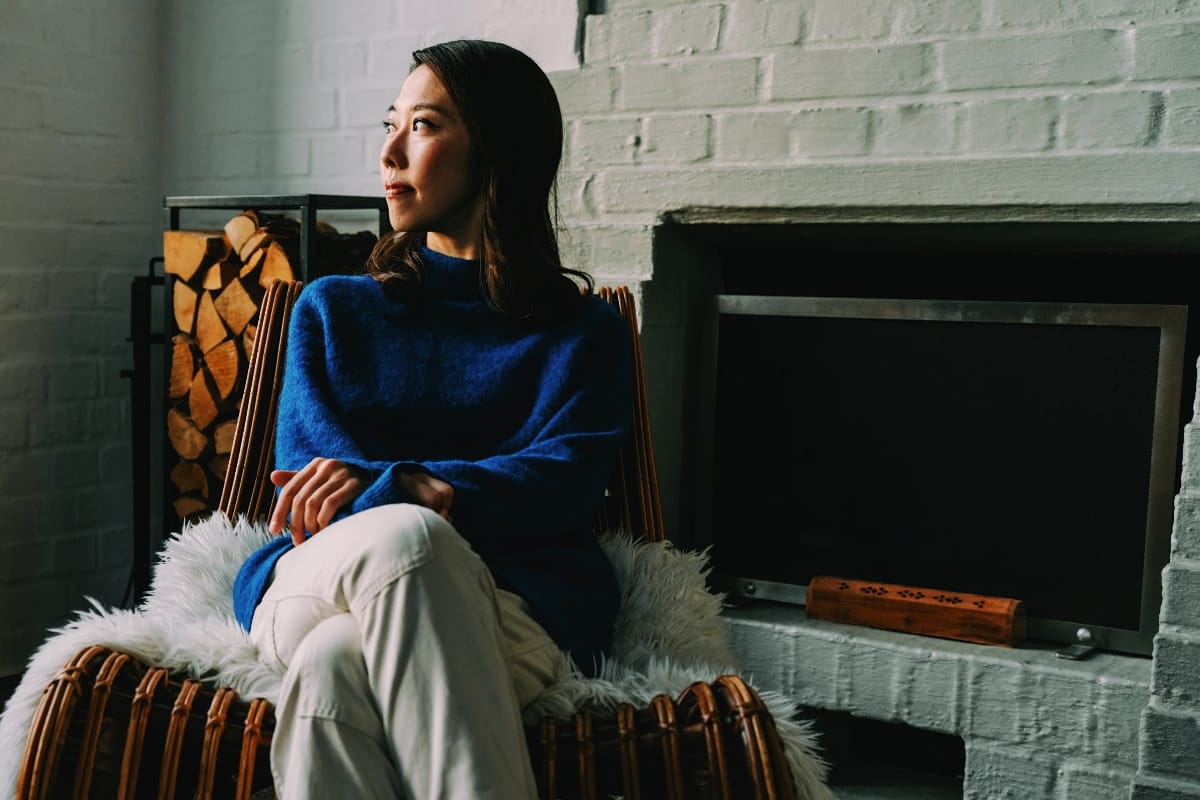 Miho Hazama sits in a wicker chair beside a white brick fireplace in a blue sweater and white trousers, gazing off to one side with stacked firewood behind her.