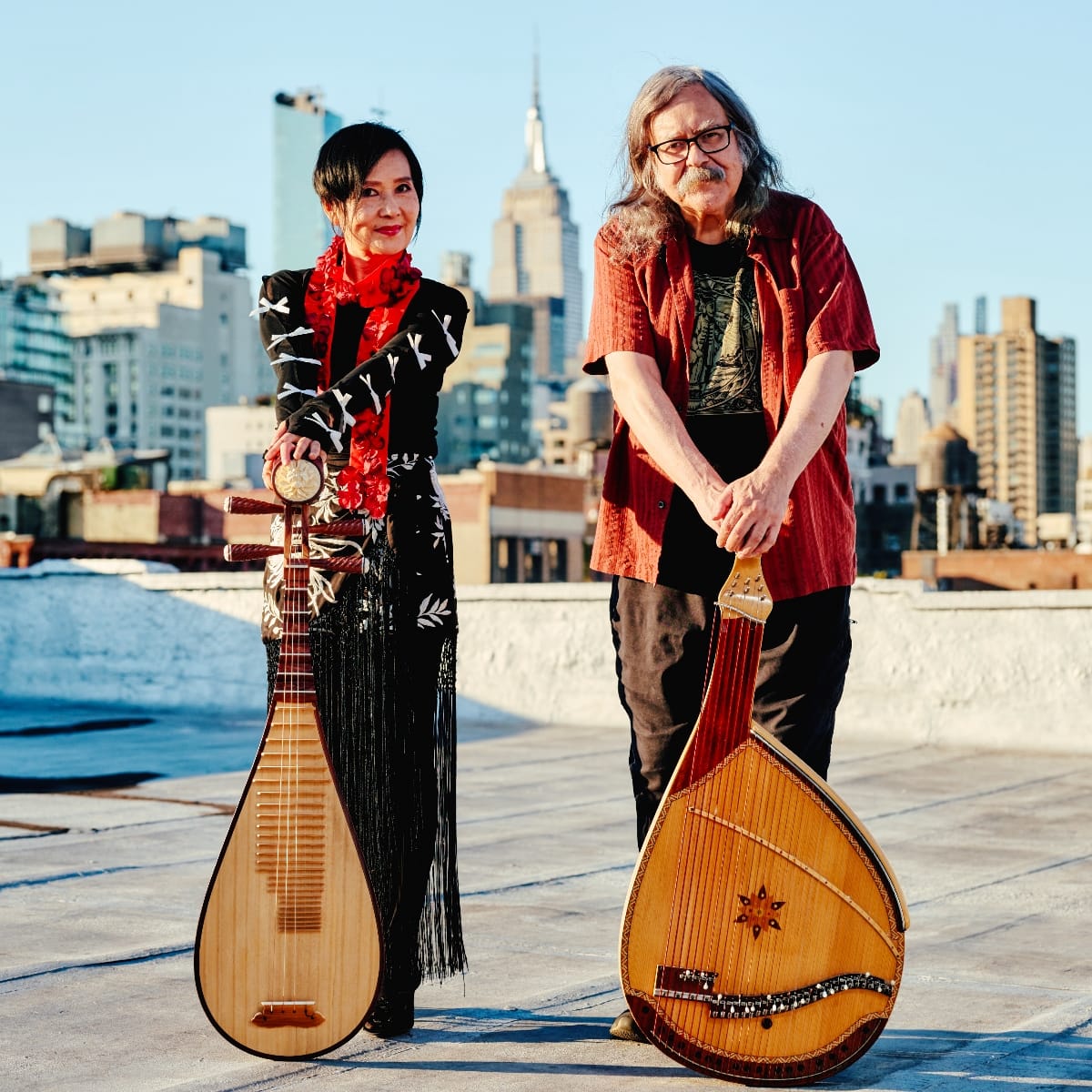 Min Xiao-Fen and Julian Kytasty stand on a rooftop with the New York City skyline behind them, each holding their instruments—a pipa and a bandura. Photo by Pavlo Terekhov. Photo by Pavlo Terekhov.