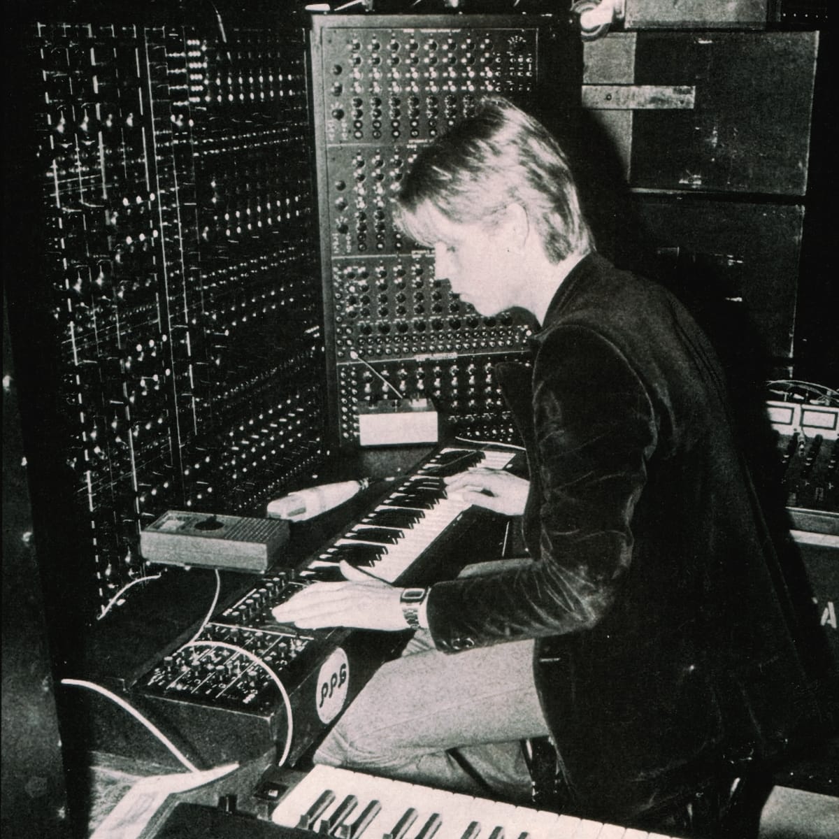 A black-and-white vintage photo of a long-haired man (Peter Baumann) playing keyboards, surrounded by towering banks of modular synthesizers and patch cables.