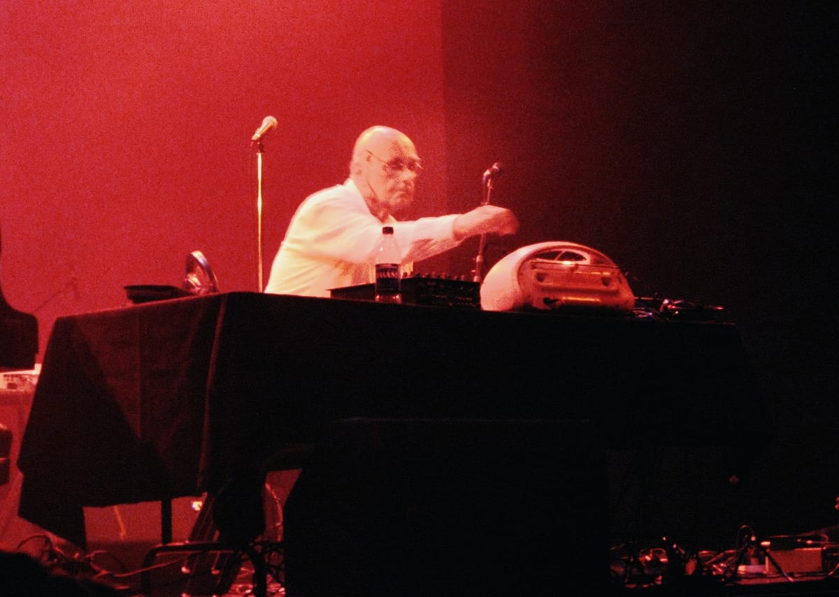 Hans-Joachim Roedelius leans over a table of electronic equipment bathed in deep red stage light during his solo performance at Moogfest, 2011. Photo by Bill Kopp.
