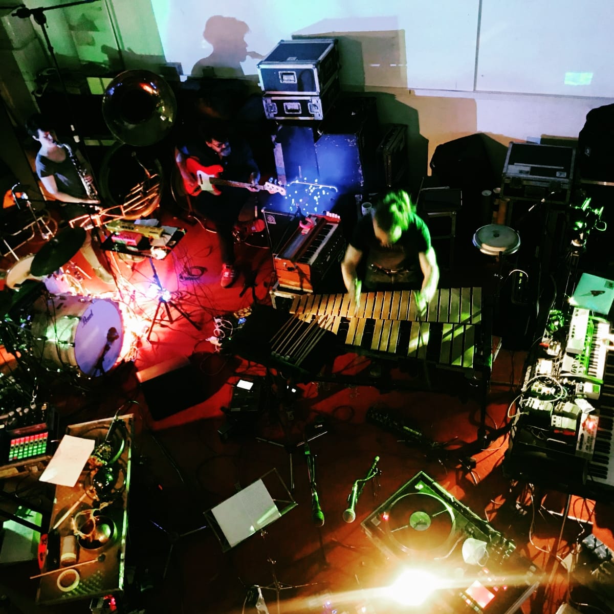An overhead view of The Notwist performing amid a dense spread of instruments and electronics, including marimba, drums, guitar, sousaphone, and modular synthesizers, bathed in red and green stage light.