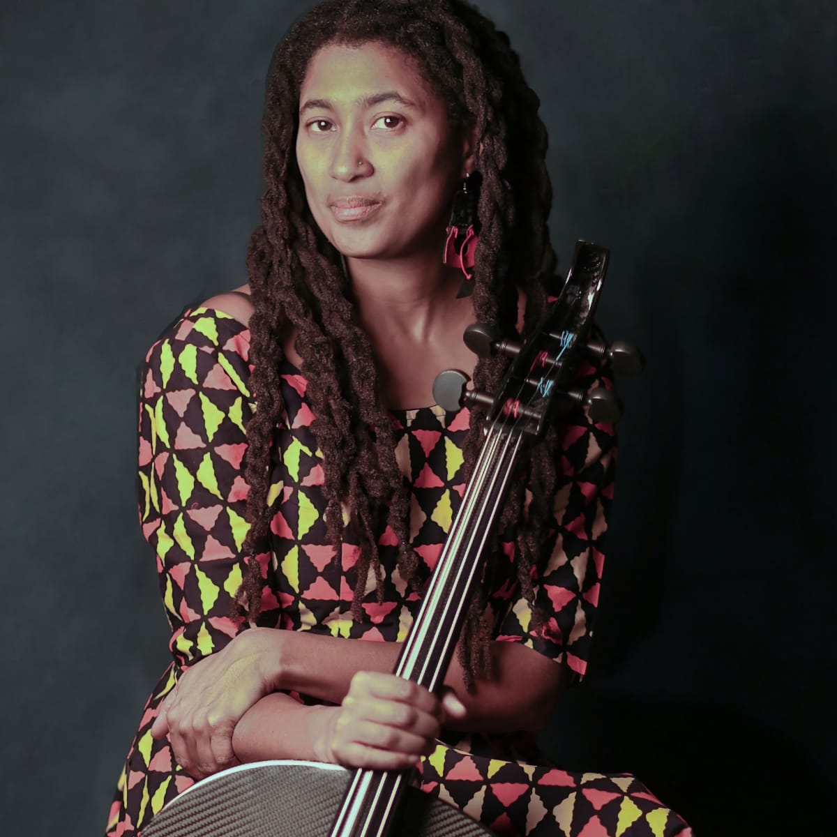 Tomeka Reid sits holding a cello in a bold geometric-patterned outfit against a dark grey background, looking directly at the camera. Photo by Michael Jackson.